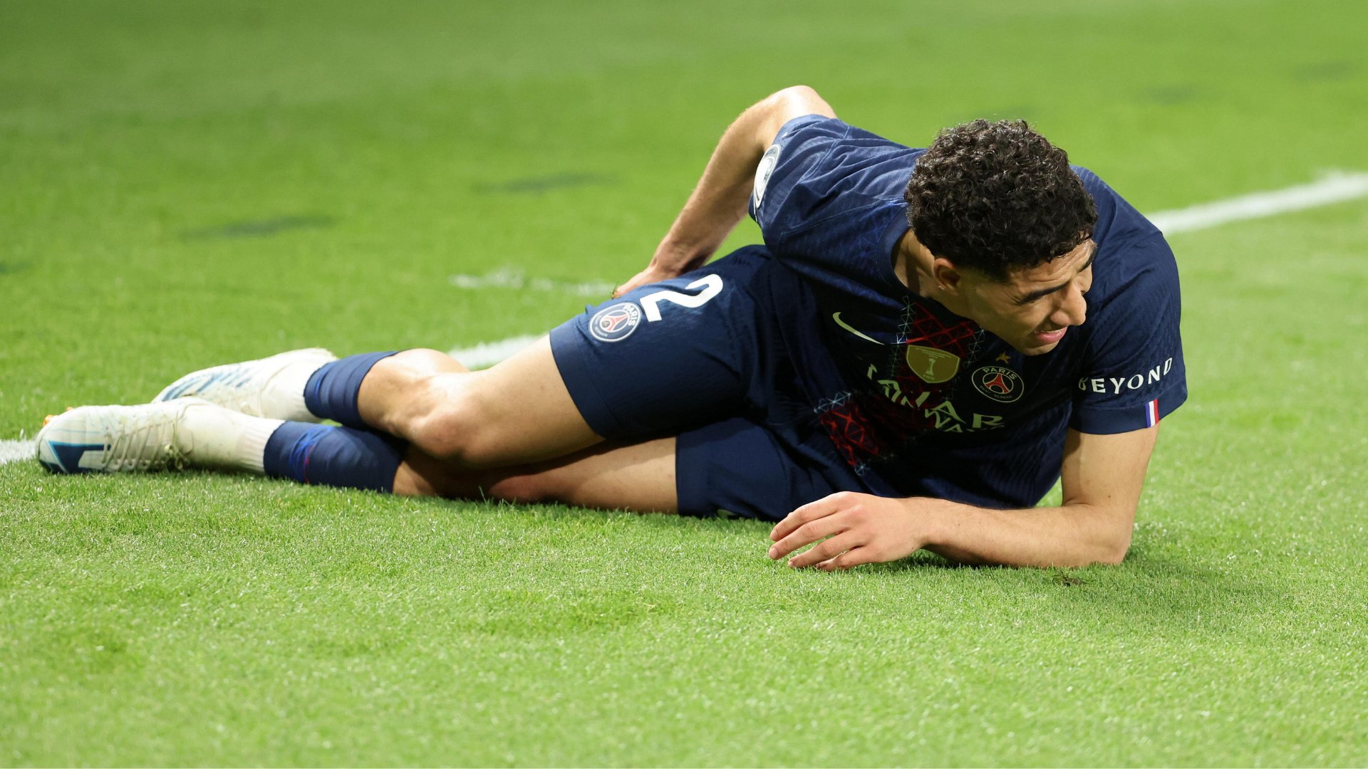 A soccer player in a dark blue Paris Saint-Germain kit lies on a green grass pitch, clutching his leg in apparent pain.
