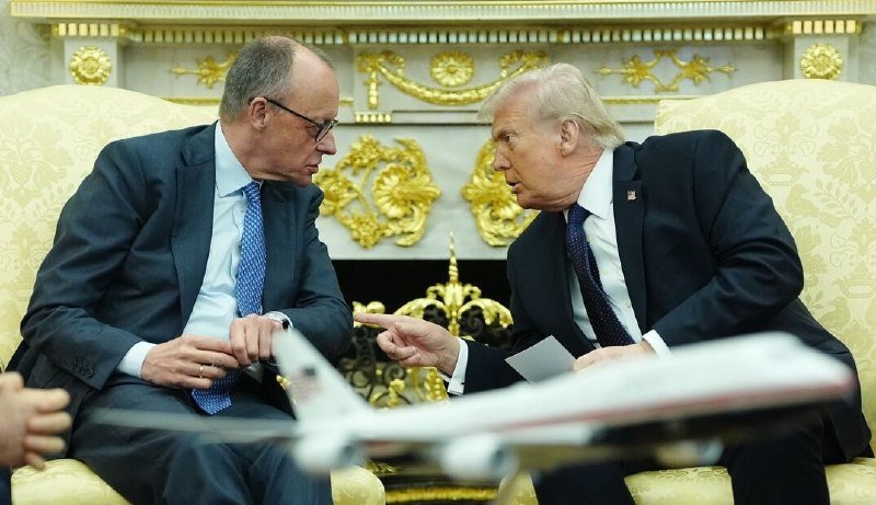 Two men in suits sit in ornate gold-trimmed chairs in an opulent room, with one gesturing toward the other across a model airplane on the table.