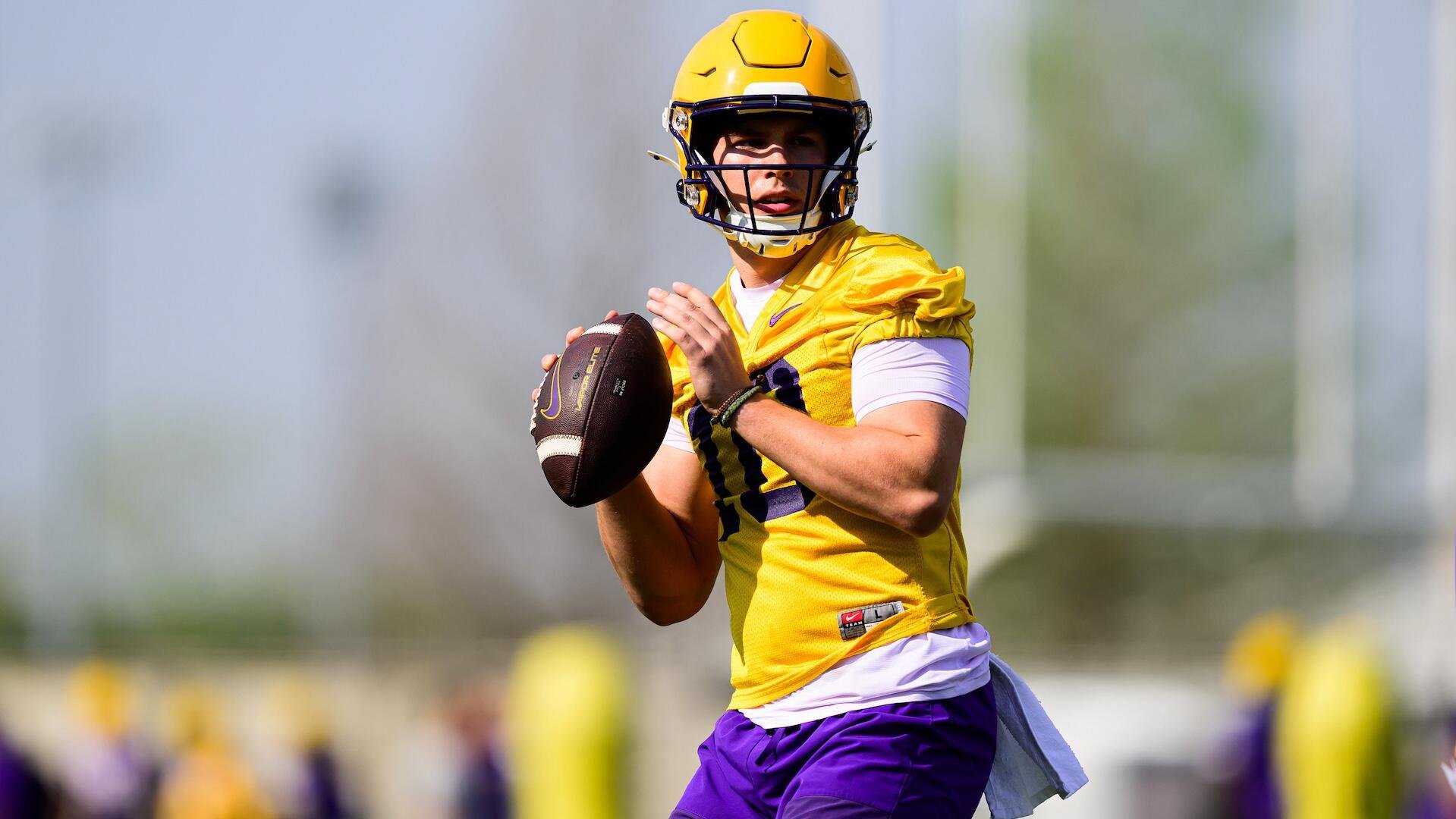 A football player in a yellow and purple uniform holds a football in throwing position during an outdoor practice.