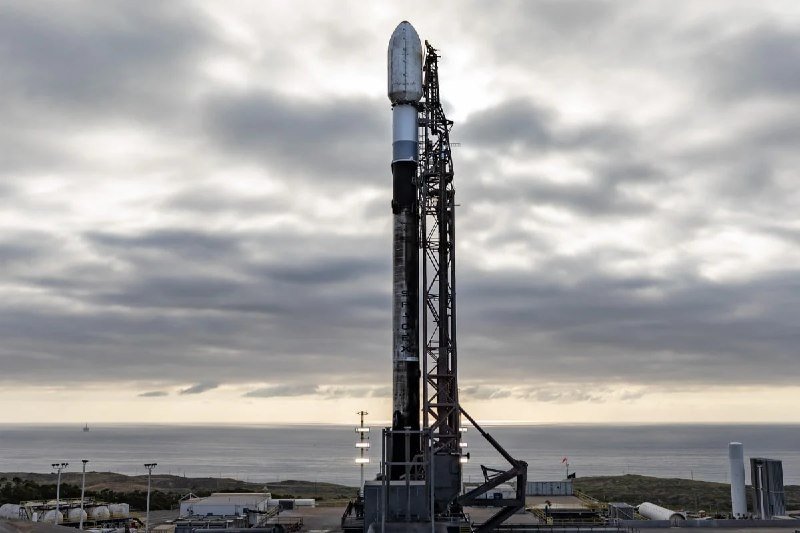 A white and black rocket marked "SPACEX" stands vertically on a launch pad beside an ocean, under a cloudy sky, supported by a tall metal gantry.