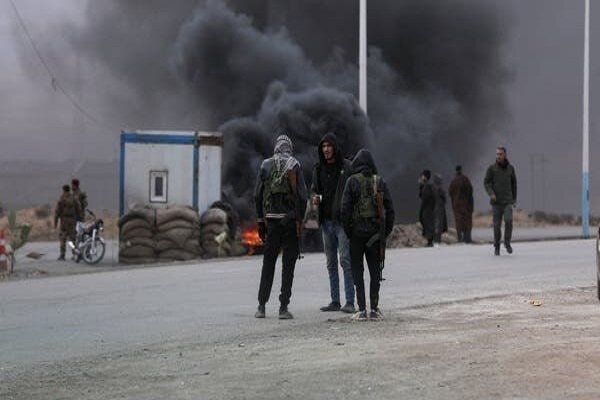 Masked individuals stand near a burning barricade as thick black smoke billows into the sky along a paved road.