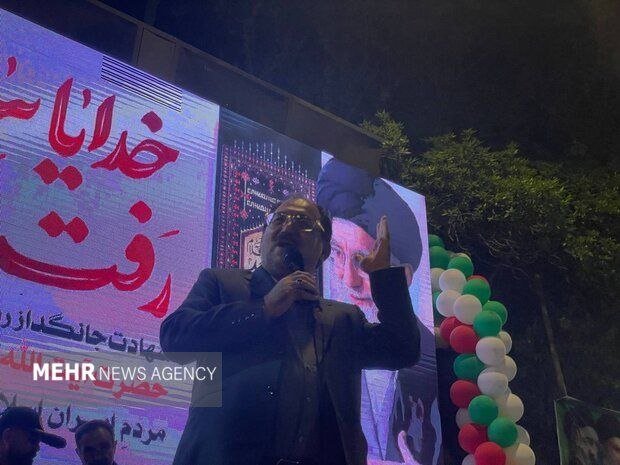 A man in a dark suit speaks into a microphone at a nighttime outdoor event, standing before a large screen displaying Persian text and a portrait, with a cluster of red, white, and green balloons nearby.