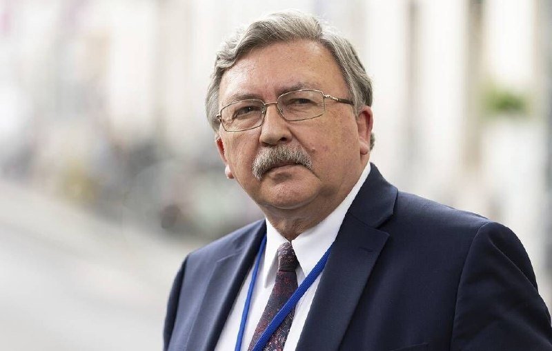 A gray-haired man with glasses and a mustache, wearing a dark suit, white shirt, patterned tie, and blue lanyard, stands outdoors with a blurred street background.