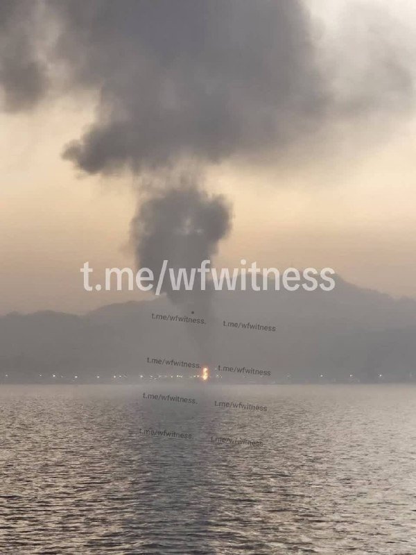 A large plume of dark smoke rises above a body of water with mountains in the background, overlaid with "t.me/wfwitness" text watermarks.