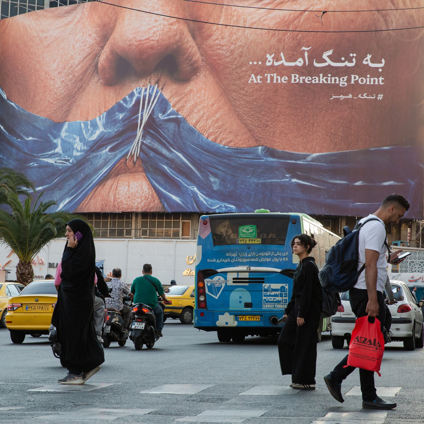 Pedestrians cross a busy street as taxis, motorcycles, and a blue city bus travel past a large billboard displaying a billboard reading "At The Breaking Point."