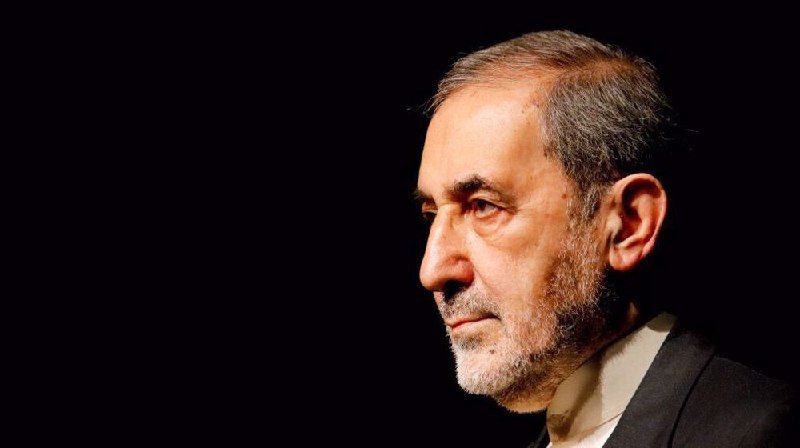 A gray-haired, bearded man in a dark suit stands in profile against a black background.