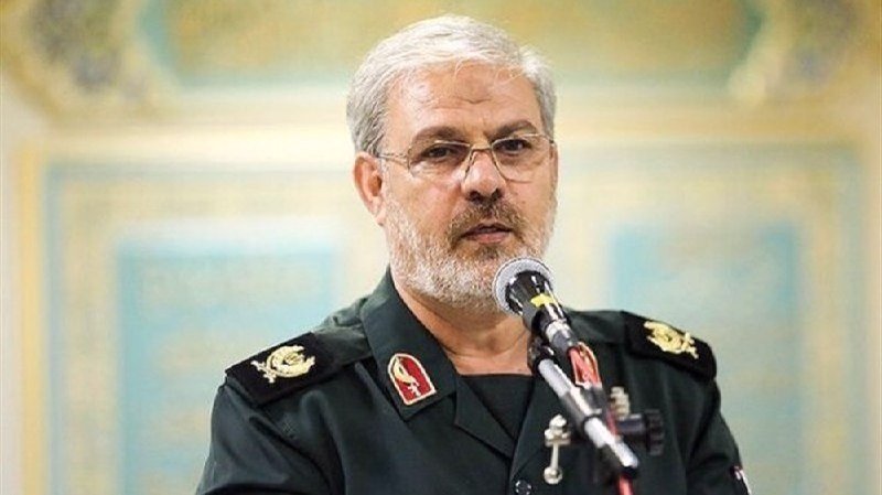 A gray-bearded man in a green military uniform with insignia speaks into a microphone.