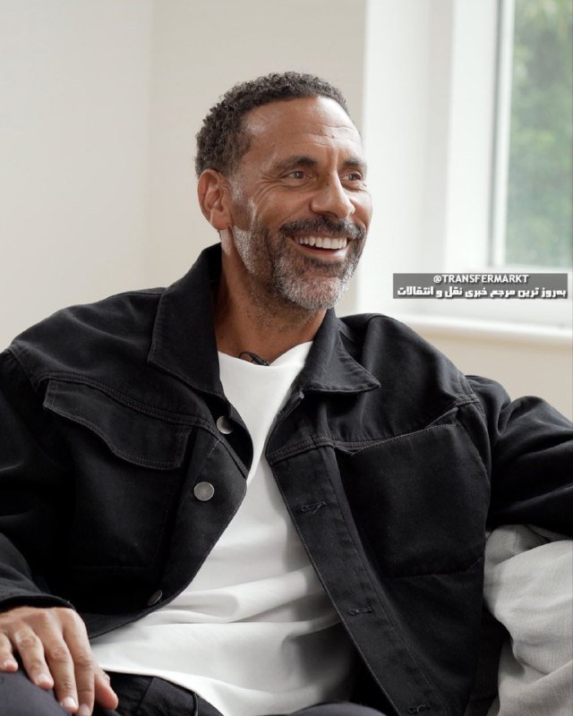 A smiling man with a gray beard, wearing a black denim jacket over a white shirt, sits indoors near a window, looking off-camera.