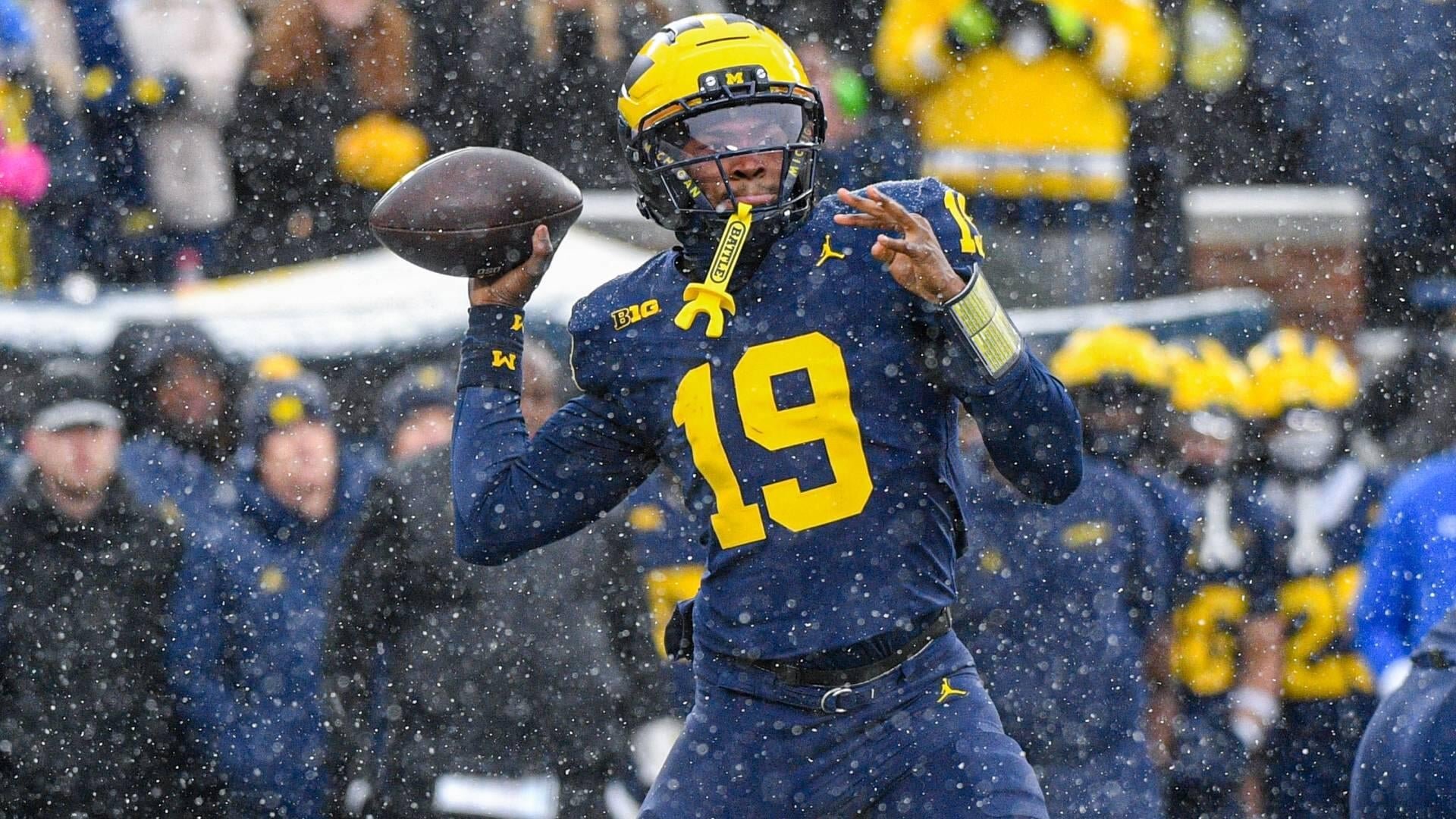 A football player wearing a navy blue and maize #19 Michigan jersey and yellow helmet throws a football in falling snow during a game.