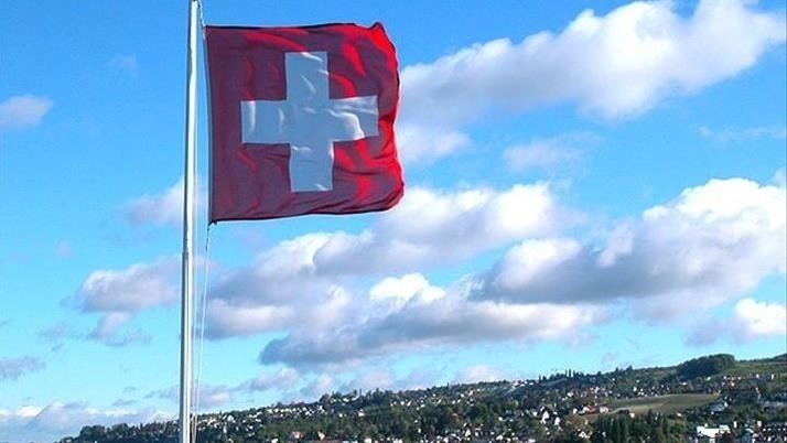 A red flag with a white cross waves on a flagpole against a partly cloudy sky, with a green hillside town visible in the background.