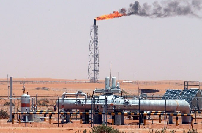 A gas flare burns atop a tall tower at an industrial facility in a desert landscape, with large pipelines and processing equipment in the foreground.