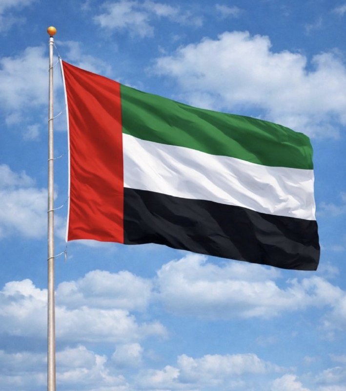 A flag with a red vertical stripe and green, white, and black horizontal stripes waves on a flagpole against a blue sky with white clouds.