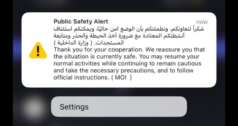 A smartphone screen displays a "Public Safety Alert" notification in Arabic and English, featuring a yellow warning triangle, with a "Settings" button visible below.