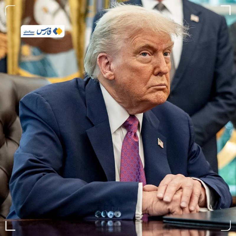 A man with light blonde hair wearing a navy suit, white shirt, and patterned pink tie sits with folded hands at a polished wooden desk.