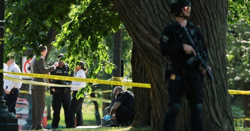 Police officers and officials stand behind yellow crime-scene tape near large trees in a park, while an armed officer patrols in the foreground.