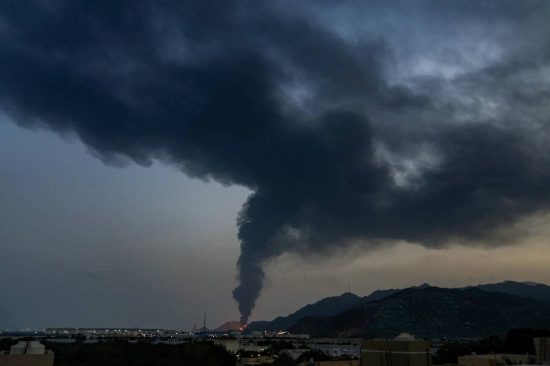 A massive plume of dark smoke rises from a fire near an industrial area at dusk, silhouetted against mountains and city lights in the foreground.