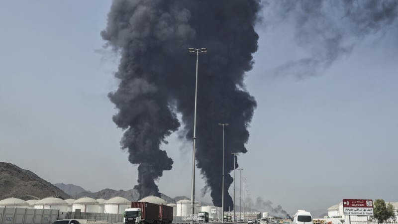 Thick black smoke billows into the sky from an industrial facility with white storage tanks, set against a backdrop of arid mountains.
