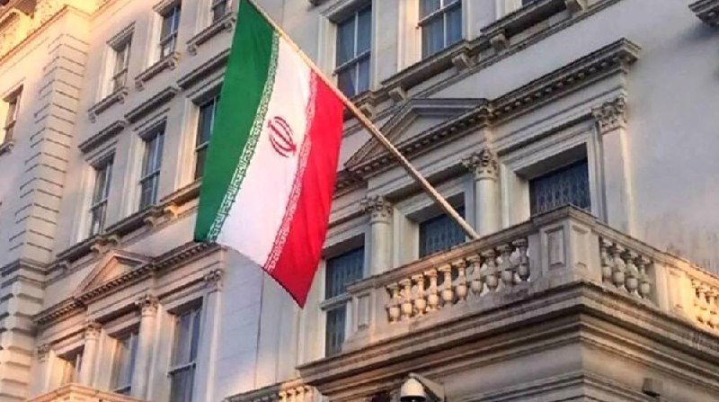 An Iranian flag hangs from a flagpole mounted on the facade of an ornate stone building with classical architectural details.