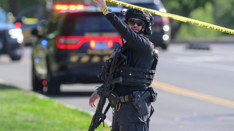 A heavily armed officer in tactical gear and a helmet stands on a roadway, gesturing with one arm raised behind yellow "POLICE LINE DO NOT CROSS" tape, with emergency vehicles visible in the background.