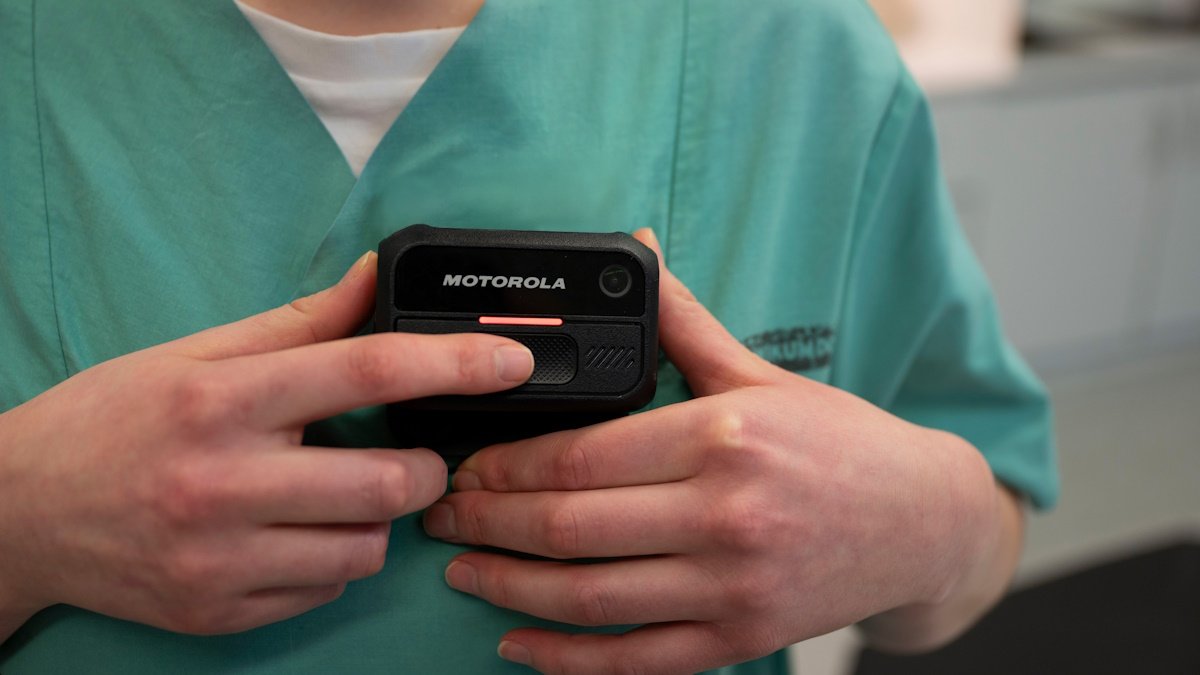 A person in green scrubs holds a black Motorola body-worn camera against their chest, pressing a button on its front.