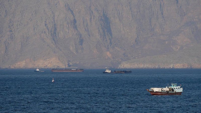 Cargo ships and a small red-and-white boat float on blue water against a steep, rocky mountain backdrop.