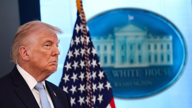 A blonde-haired man in a dark suit stands beside an American flag, with a White House logo displayed on a screen behind him.