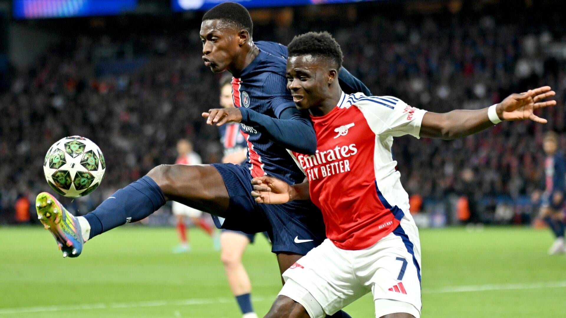 Two soccer players—one in a dark blue kit, the other in a red-and-white "Emirates Fly Better" jersey—compete for the ball during a match in a crowded stadium.