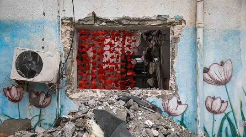 A damaged building wall features a broken window opening filled with red debris, surrounded by a white air conditioning unit and painted pink flowers on light blue walls.