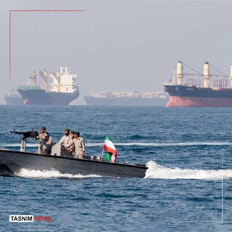 Four armed personnel in camouflage navigate a small speedboat flying a green-white-red flag across open water, with large cargo ships visible in the hazy background.
