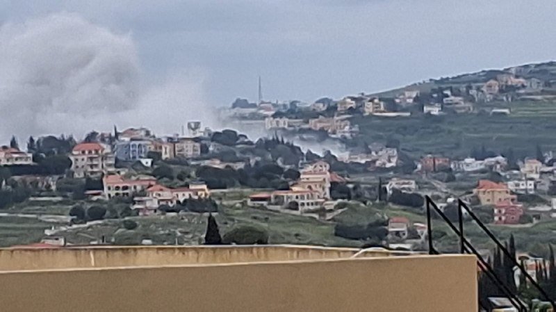 A plume of white smoke rises from a hillside residential area with scattered houses under a cloudy sky.
