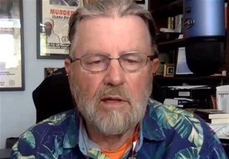 A bearded man wearing glasses and a tropical-print shirt speaks in an office setting with framed posters, bookshelves, and a blue object visible in the background.