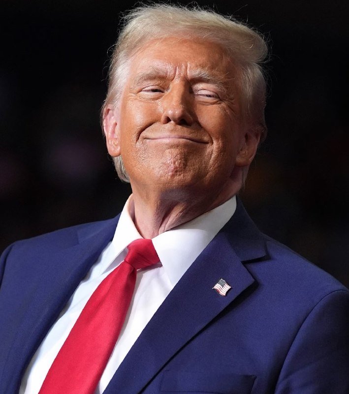 A man with light blonde hair, wearing a navy blue suit, white shirt, and red tie with an American flag lapel pin, smiles against a dark background.