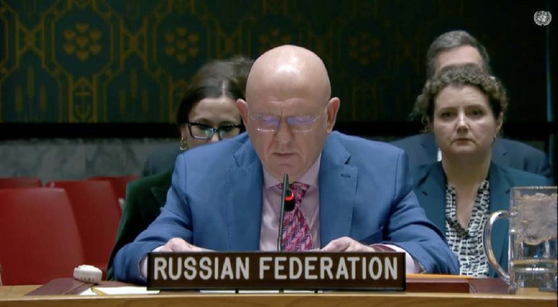 A bald man in a blue suit speaks into a microphone behind a "RUSSIAN FEDERATION" placard at a formal assembly, with seated attendees visible behind him.