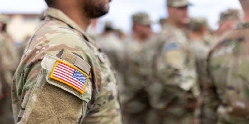 Soldiers in camouflage uniforms stand in formation outdoors, with an American flag patch visible on the foreground soldier's sleeve.