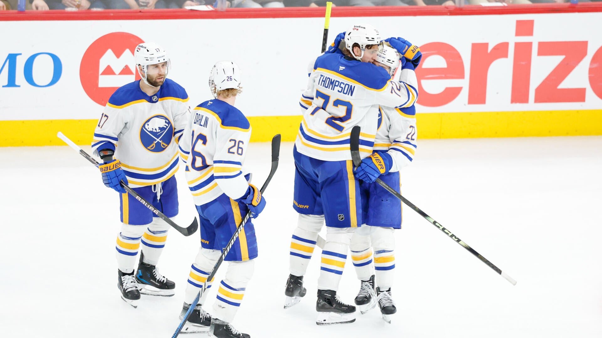 Buffalo Sabres hockey players in white and blue uniforms embrace on the ice in celebration, with teammates skating toward them, in front of a "Verizon" advertisement on the boards.