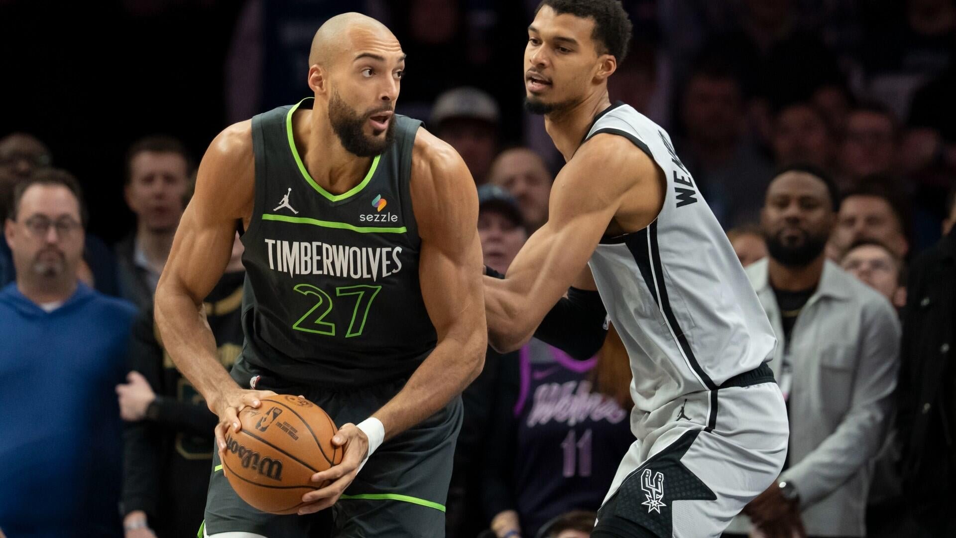 A Minnesota Timberwolves player wearing jersey number 27 holds the basketball with both hands while being guarded by a San Antonio Spurs player in a white uniform during a game.
