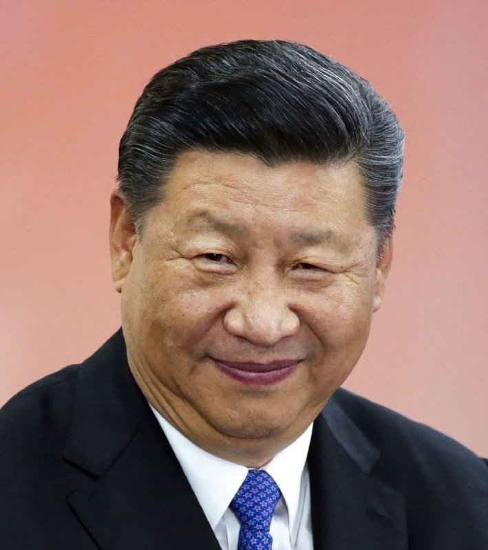 A man with dark, graying hair in a dark suit and blue patterned tie smiles against a solid pinkish background.