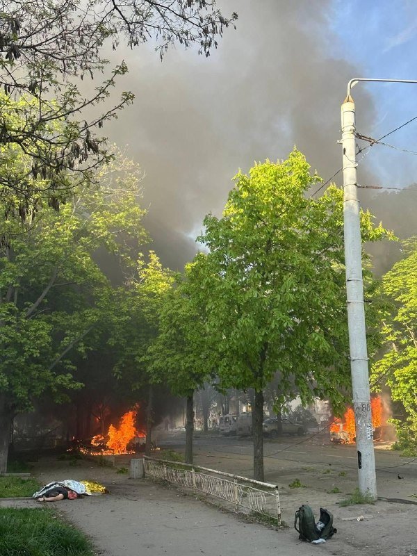 Orange flames and thick gray smoke rise along a tree-lined street, with a person lying on the sidewalk and a backpack nearby.