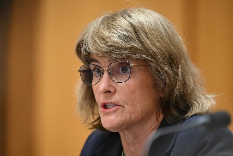 A blonde woman wearing glasses and a dark blazer speaks into a microphone against a wooden panel background.