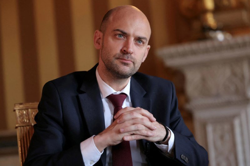 A bald, bearded man in a dark suit and burgundy tie sits with hands clasped in an ornate room.