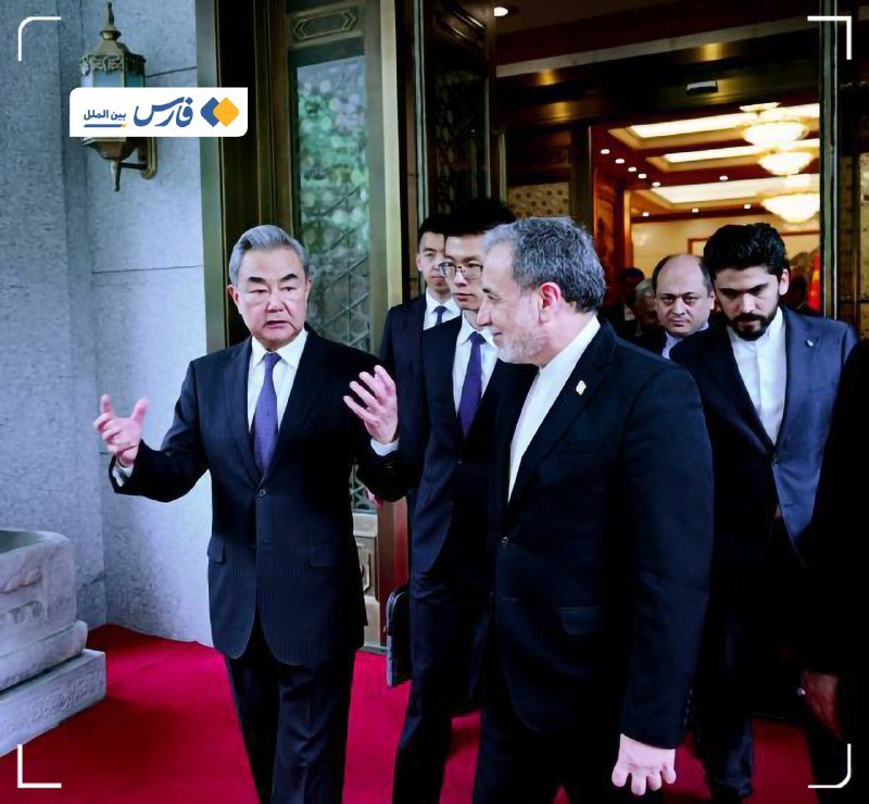 A group of men in dark suits walk together through an ornate doorway onto a red carpet, with one gray-haired man gesturing with his hand as he speaks.