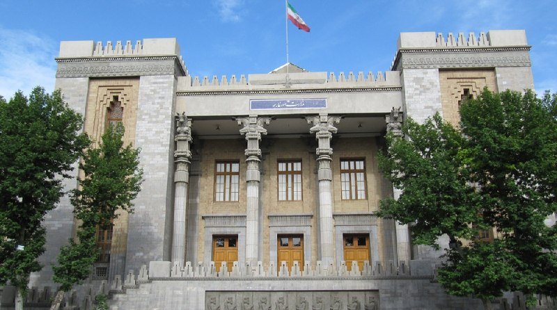 A grand stone building with classical columns, arched doorways, and an Iranian flag flying atop, flanked by green trees under a blue sky.