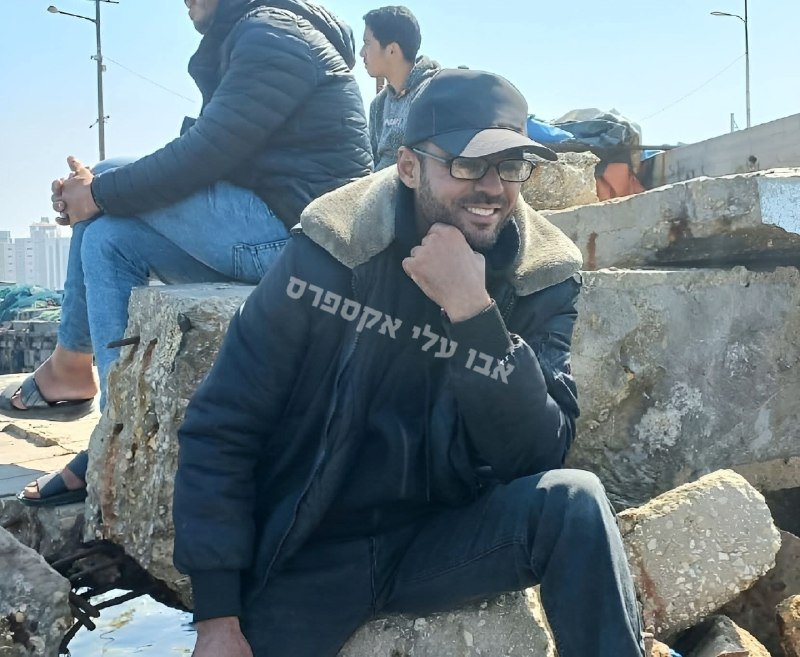 A smiling bearded man wearing a cap, glasses, and a hooded jacket sits on concrete rubble near water, with other people and debris visible behind him.