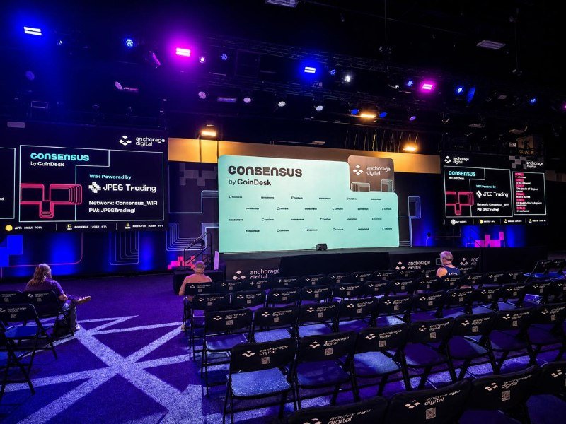 Empty rows of chairs face a brightly lit stage displaying "CONSENSUS by CoinDesk" signage with "WiFi Powered by JPEG Trading" advertisements under purple and blue lighting.