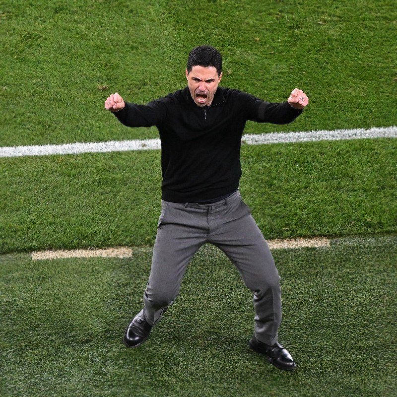 A man in a black sweater and gray trousers stands on a grass field with clenched fists raised and mouth open in a shout.