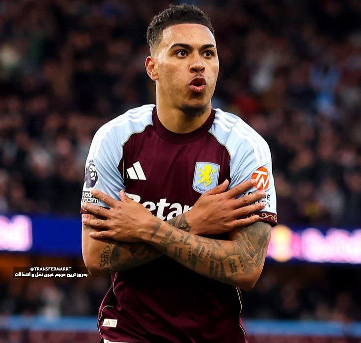 A soccer player in a claret and light blue Aston Villa jersey crosses his arms over his chest during a match in front of a crowded stadium.