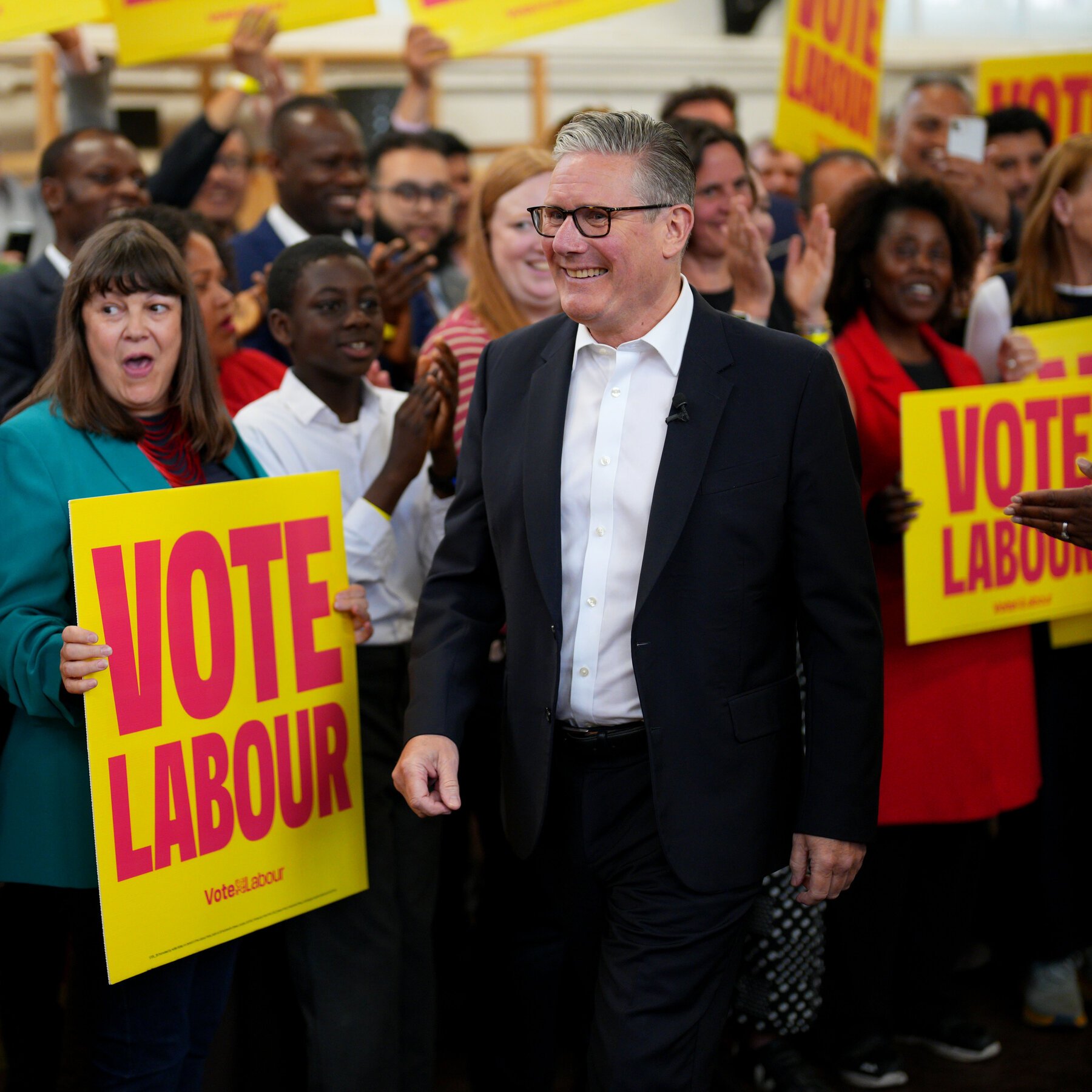 A smiling man in a dark suit and glasses walks through a cheering crowd holding yellow and red "VOTE LABOUR" signs at an indoor political campaign event.