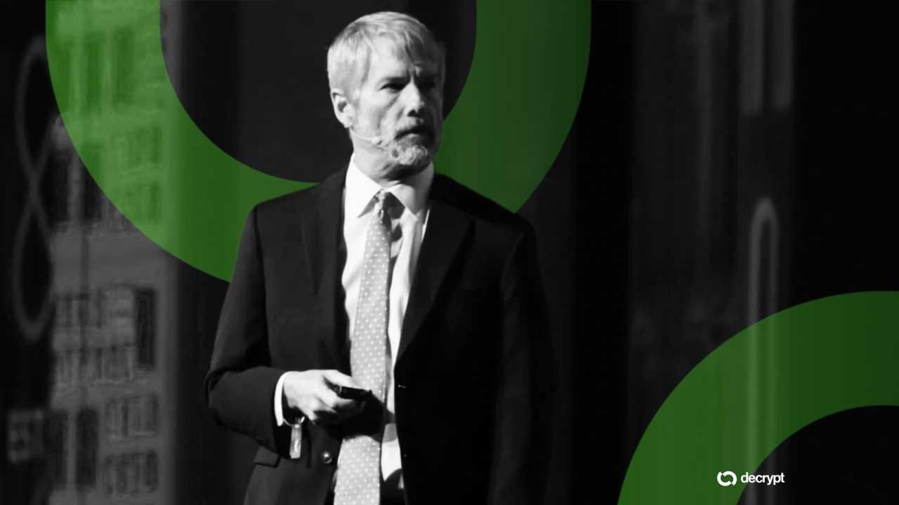 A gray-haired man in a suit and tie holds a presentation remote while speaking on stage, overlaid with green circular graphics and a "decrypt" logo.