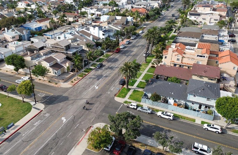 An aerial view shows a suburban intersection with parked cars, palm trees, and residential homes lining the streets.