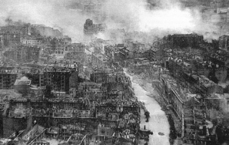 Black-and-white aerial photograph showing a heavily destroyed urban area with ruined buildings and smoke rising across the cityscape.